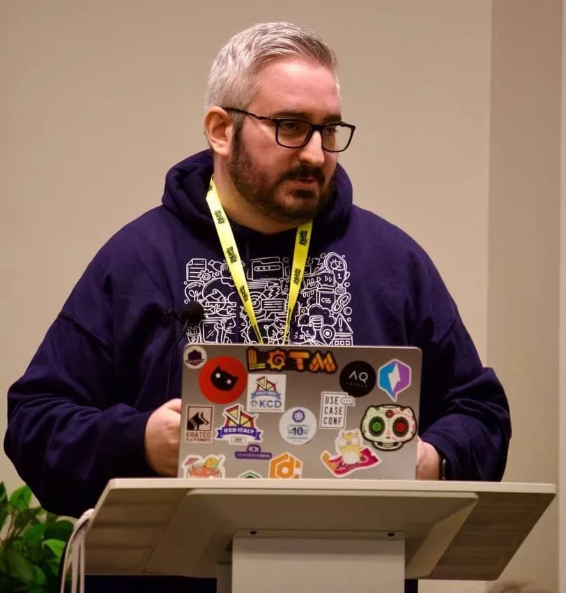 Davide at a lectern, laptop covered in community stickers